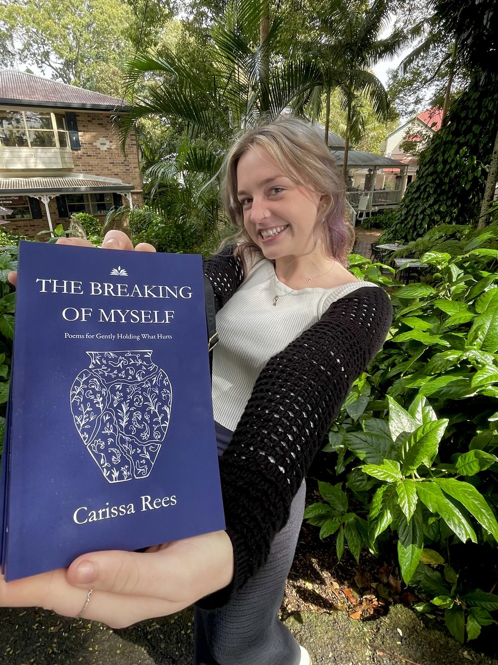 Carissa Rees smiling in a leafy garden, holding up a copy of The Breaking of Myself.